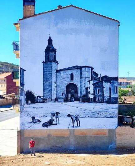 Streetartwall. On the outer wall of a three-story house is sprayed / painted mural of a village view with church. In the colors blue, white and black, the square is depicted as it looked in the sixties. A church with with tower and large gate is in the center. Next to it two other typical Spanish buildings. In addition, a large empty square and in the foreground 4 dogs are standing and lying on a path. On the photo the artist stands proudly in front of his mural. Another one has already been started.