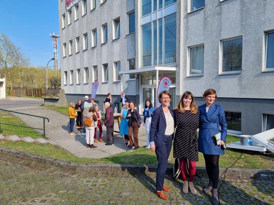 Drei Frauen vorm Gebäude im Hintergrund Menschen im Gespräch.