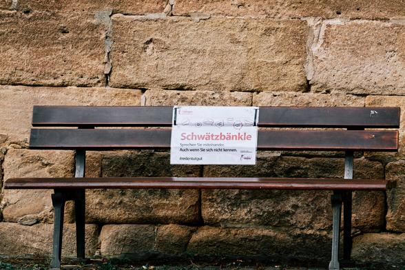 Auf dem Bild ist eine leere Sitzbank aus braunem Holz vor einer Kirchenmauer zu sehen, auf der ein eher behelfsmäßiges Schild – ein folierter Ausdruck – mit folgendem Text zu sehen ist:
„Schwätzbänkle – Sprechen Sie miteinander. Auch wenn Sie sich nicht kennen.“ Plus dem Schlagwort, einem Hashtag #RedenTutGut