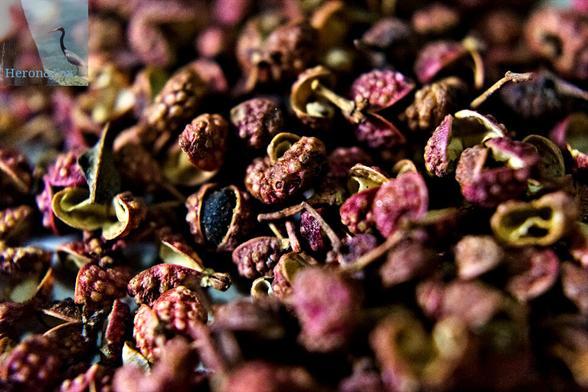 A close-in, macro photograph of Sichuan peppercorns, which appear as small, black spheres, coated in a thin shell of material colored in reds, purples, pinks, yellows, and golds. It has a mildly knobby texture and peels away from the dried, black berry.
The image appears to depict part of a tall slop of such peppercorns.