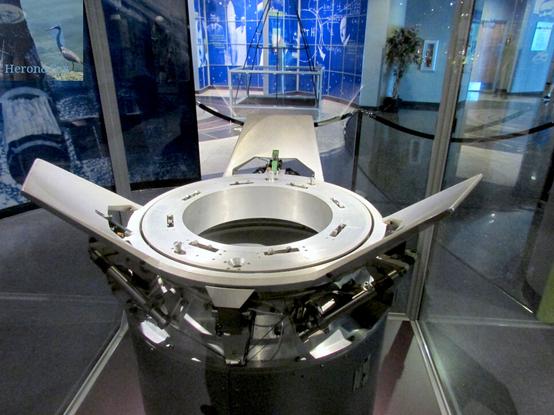 A photograph of an Androgynous Peripheral Attach System, or APAS, docking ring on display.
The docking ring is made of silver metal and three “petals” project up and out from its edges. It sits on a mechanical base that sits on a black pedestal.
