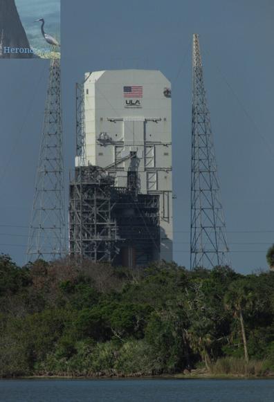 A photograph of Space Launch Complex 37B. At the bottom of the frame is a sliver of water, and some scrub land above that. The background is dominated by an imposing, rectangular white tower. There is a US flag and a ULA logo on top. The top half of the building is closed in by doors that look like they slide off to the sides. The bottom is open and a single orange, cylindrical Delta 4 rocket core can be seen.
There is a gray, truss tower on the left hand side of the building with connections that reach from the truss to the rocket.
On either side of the white building are tall truss towers which serve as lighting deflectors.