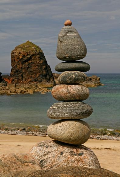 Art imitating nature. We are on a beach looking out to the sea. In the distance is a sea stack, in the foreground is a stack of vertically balance rocks. One vaguely resembles the other