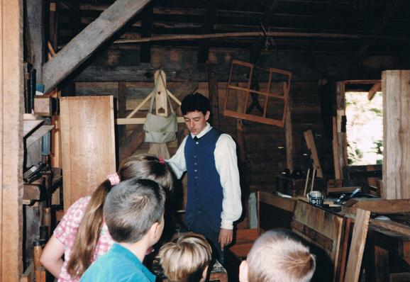 A photo of me taken in the early 1990s when I worked as an Apprentice Carpenter/Joiner with the Colonial Williamsburg Foundation in Williamsburg, VA. In this image I am teaching some children about how to properly plane a board flat and square. I am, of course, wearing an 18th century tradesmen’s costume.