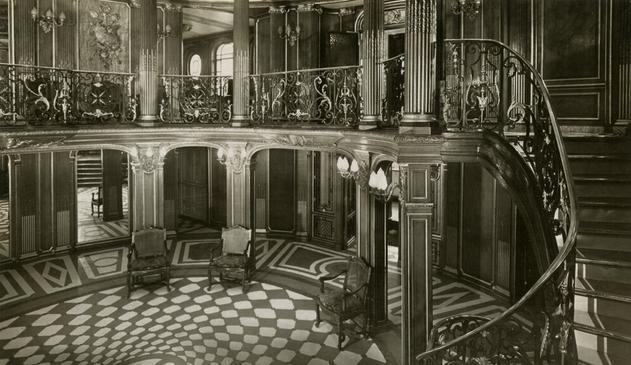 Black and white photo of the grand staircase and foyer  of the SS France, April 1912. This is a two-deck space with ornate columns, paneled pillars on the ground level, and a terrazzo floor with an oval-shaped geometric design. The décor is Baroque and is clearly a French style.