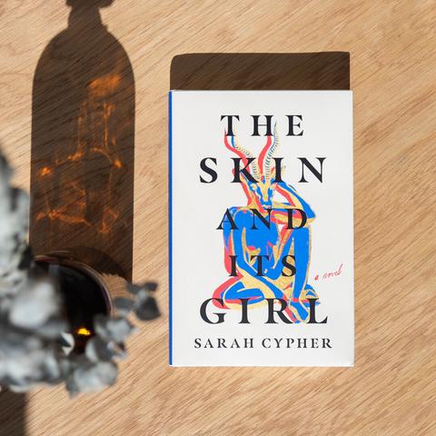 The cover of THE SKIN AND ITS GIRL depicts the title in large black words arranged vertically over the illustration of a cobalt-blue female figure who has a striking gazelle head. The book sits on a hardwood surface next to the shimmering shadow of a glass bottle.