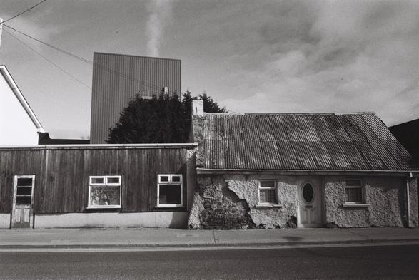 a black and white wide angle film photograph of a street in Kanturk, Co. Cork, Ireland. There are old houses in the front, with a large industrial building looming in the background, with a pillar of steam rising