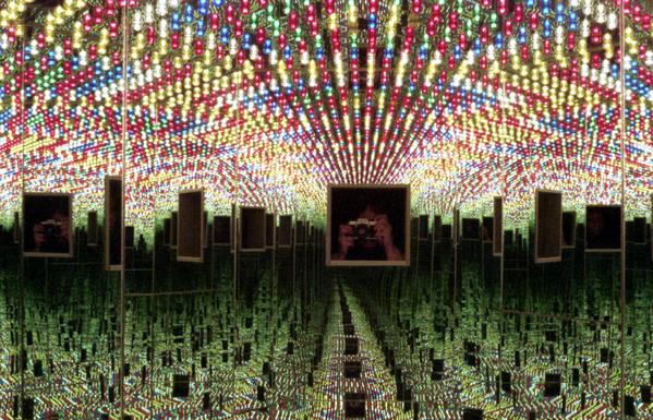 A person aims a camera into a small enclosure covered in mirrors and multicolored lights.