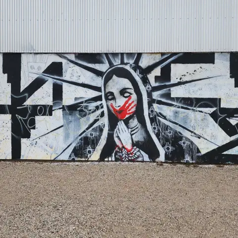 broader frame of black and white mural of Tonantzin with a red handprint over mouth, hands bound with rope in traditional prayer position, and Zia symbols on robe instead of stars