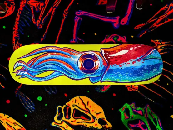 A brightly coloured skateboard painted with a Japanese Firefly Squid. Pictured on top of dinosaur bone carpet.