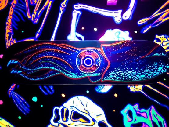 A brightly coloured skateboard painted with a Japanese Firefly Squid. Pictured on top of dinosaur bone carpet. A UV flashlight shines on the skateboard to show off the UV reactive paint.