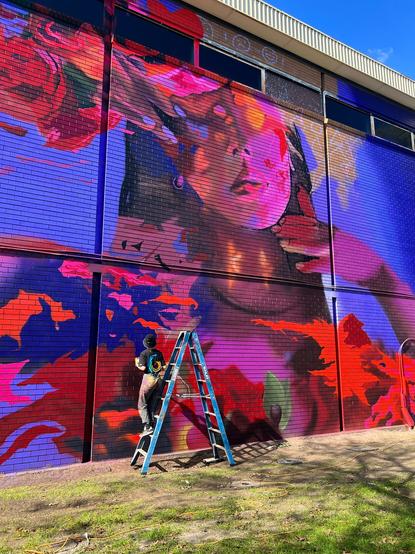 The Melbourne artist (Juzpop) on a small ladder, poised to place brush to the wall, one third up from the ground. 

Painting is partially complete; I’d suggest half-way done, with the basic layers applied on a brick wall about 20-25 metres in length and 7-8 metres high. This building is an eye sore, an old brown brick telephone exchange building, I know it well. 

The main feature of this creation is a young woman’s face, looking confident, determined and empowered, although a touch of softness and vulnerability too (my interpretation)… most likely early to mid-twenties, pretty, tanned complexion, black hair and red lips (face is not complete), with her hands framing her face, blue fingernail polish on right hand (left not yet complete). 

Her face is surrounded by a sea of vibrant blues, purples, reds, pinks and subtle oranges which are to become leaves and foliage of some sort amongst other things I think… perhaps in bright and dynamic, autumn colours going by the aesthetics thus far! Also roses or some other flower forms are perhaps in the making too. 

I can see this coming along nicely, with shading and more depth and detail to come. This will look absolutely breathtaking once complete. Feminine beauty and strength at its finest. Just wow.