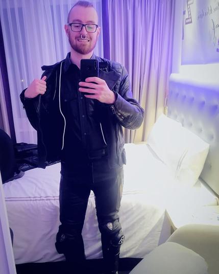 A man with a septum piercing dressed in black leather biker pants, leather shirt, vest and jacket and leather boots taking a selfie in a hotel mirror.