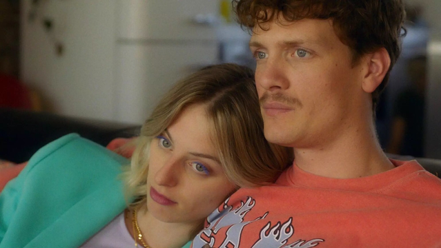 Tessa (links, Claire Bender) en Paul (rechts, Martijn Lakemeier) zitten op een bank binnen. Het hoofd van Tessa leunt op de schouder van Paul. Beiden kijken met een onbestemde blik naar buiten. Paul draagt en oranje seater, Tessa een mintgroene. Beiden zijn jonge, witte mensen.