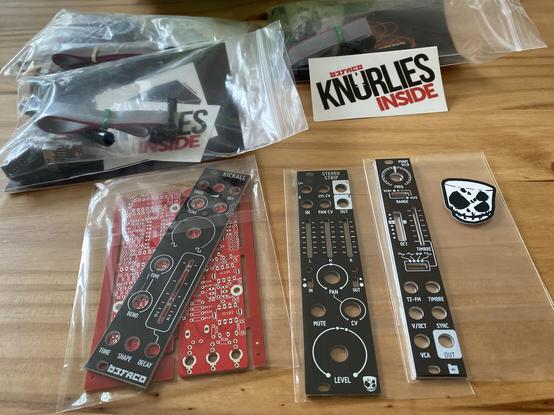 Front panels of Befaco Eurorack modules, from left to right: Kickall, Stereo Strip, and Pony VCO. A sticker of Divkid logo is to the right. Behind the panels there are three plastic cables with ribbon cables and electronic components in them. "Befaco Knurlies Inside" sticker is on one of the bags.