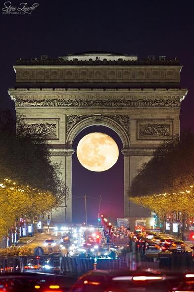 A nearly full Moon is seen through the famous Arc de Triomphi with trees and cars lining the foreground.