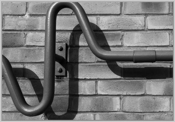An abstract black and white photograph of a enameled steel hand-rail with a decorative pair of curves in strong, angled, sunlight and a shadow as a twin falling on the backdrop of brickwork.