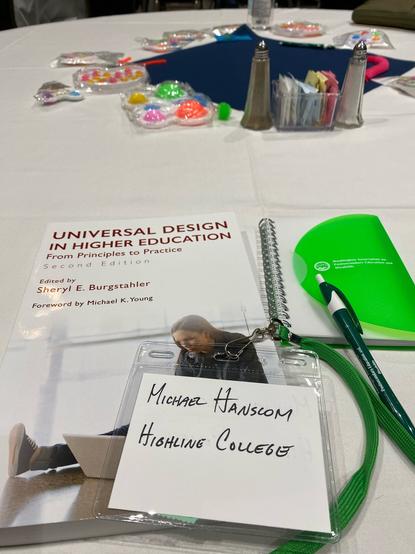 A table with a selection of fidget toys arrayed in the center. In the foreground is a handwritten conference name tag, a WAPED branded pen and small notebook, and the book _Universal Design in Higher Education: From Principles to Practice_ (2nd ed.).