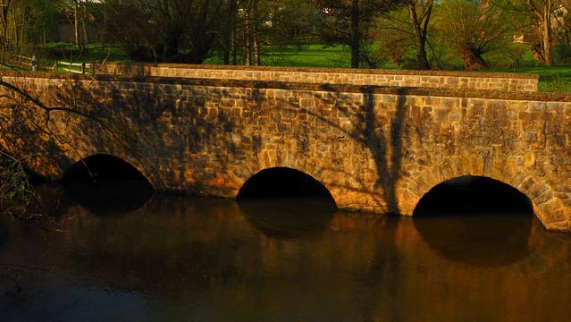 Bruchsteinbrücke mit drei Bögen über einen kleinen Fluß. Die tiefstehende Sonne lässt alles warm und golden erscheinen