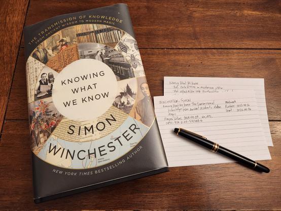 Simon Winchester's book Knowing What We Know sitting on a wooden table next to a bibliography card and some notes written on index cards along with a black Mont Blanc fountain pen with gold trim.