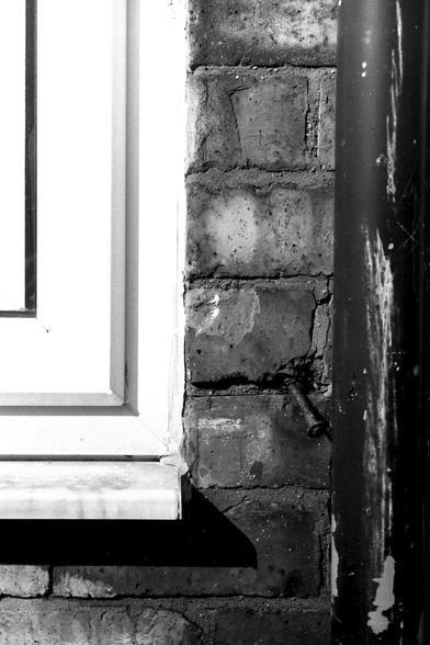 A 35mm black and white film photograph of a part of a white uPVC window, a brick wall and a downspout from my guttering showing the transitions between the three textures