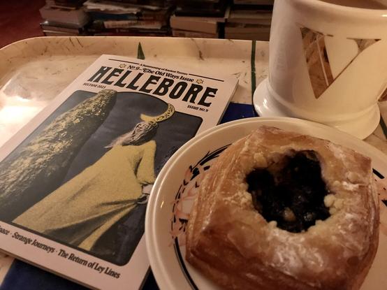 A tray holding the Hellebore zine number 9, the Old Ways issue for Beltane 2023. The cover of the zine shows a woman beside a tall old stone with a dark sky behind. Beside the zine there is a huge Danish pastry with a dark fruit filling. And behind that is a large cream mug of coffee with a gold monogram V on the outside.