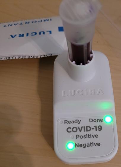 Lucira test in foreground: Electronic device showing "Done" light and "COVID-19: Negative" light. Test tube with purple fluid is in the top of the device. Background: instructional brochure with words "Lucira" and "Important" visible.