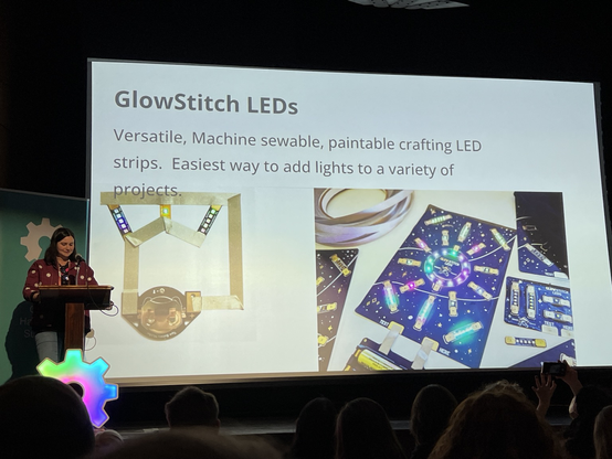 Steph stands on stage, in front of a slide showing GlowStitch LEDs – "versatile, machine sewable, paintable crafting LED strips. Easiest way to add lights to a variety of projects."