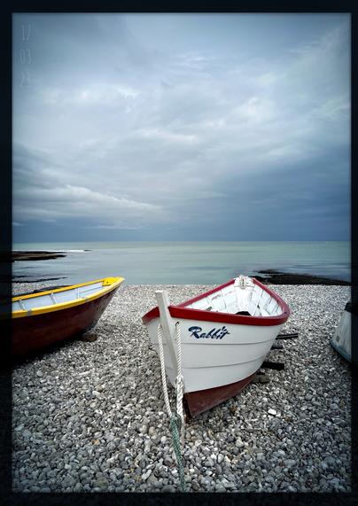 Das Foto zeigt im Vordergrund einige Holzruderboote, die auf dem Kiesstrand des kleinen Küstenortes Yport in der Normandie liegen; im Hintegrund dominiert der dunkle Wolkenhimmel über dem recht ruhigen Meer, der den Aufzug eines Gewitterschauers ankündigt; ein dunkelblauer Schriftzug "Rabbit" ziert das weissrote Ruderboot im Fokus des Bildes, wobei über dem i-Punkt zwei kleinen Ohren die Typographie ergänzen