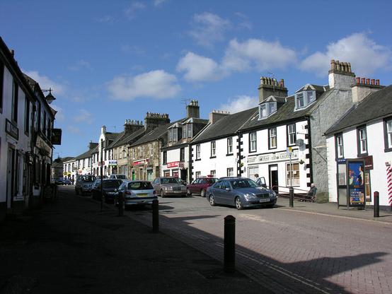 Cumbernauld village in North Lanarkshire. The image shows a view along a street in sunlight with the left-hand side in deep shadow. The buildings are two storeys high, some with dormer windows in their attics, and most are finished in white.