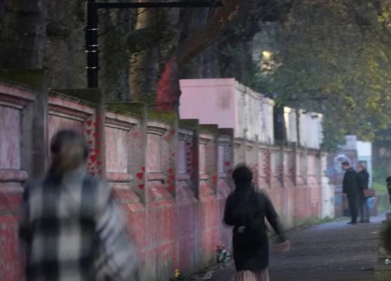 Slightly blurred figures walk past a wall whilst looking at it. The wall has many red hearts painted on it, and bunches of flowers leant against it.
