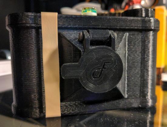Close up photo of a black, plastic pinhole camera from the front. There's a large rubber band securing the top cover. The "shutter" is simply a hinged door that you lift to make an exposure, and close when you think it's done. The exposure time can vary from a second or two up to many minutes in length, so precision in timing isn't a critical concern.