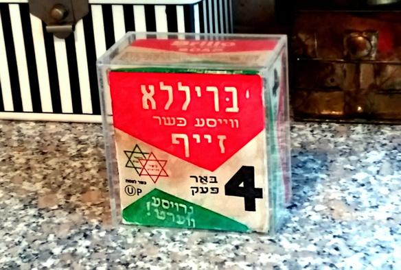 A clear plastic box containing on old cardboard box with Yiddish writing on it, two Jewish stars, an OU-Pareve symbol, and the number 4.