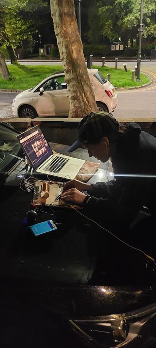 tinysound is live-testing a self built radio receiver (an Open Wave-Receiver) on the bonnet of a black car at night with a road, car and trees in the background. She is appearing live in an online workshop to build radios via a laptop which is also on the bonnet of a car. She is also wearing a metro tartan cap which she bloody loves, though is unnecessary for night time lol.