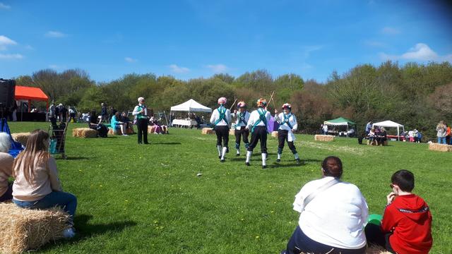Morris Dancers dance with sticks in a sunny field in front of an audience sitting on hay bales