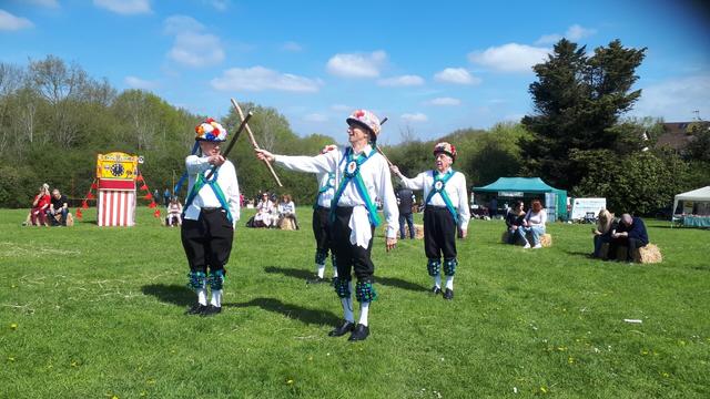 Morris Dancers finishing a dance by crossing their sticks in a sunny field in front of a Punch and Judy tent, and an audience sitting on hay bales