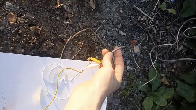 Alyssa’s open wave-receiver can be seen from above with her mounting board on some gravel and next to some stinging nettles. Alyssa’s hand is holding a wire-connection on her radio where crocodile clips are connected to her antenna which trails off to the right and into the stinging nettles.