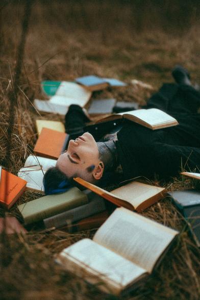 Photo of a nonbinary person lying on their back on the ground in a field of tall brown, dormant winter grass. They have fair skin, a blue & black mohawk hairstyle, and are dressed in black. Tattoos are visible on their neck and chest. An open book rests on their chest and their eyes are closed. They are surrounded by hardcover books, which are messily scattered around and under them - some open, some closed. Photo by Emma Wondra.