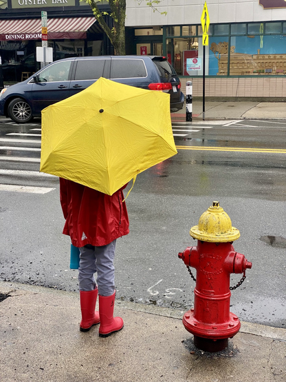A child with a red raincoat, red boots, and a yellow umbrella stands beside a matching red-and-yellow hydrant on a city street.