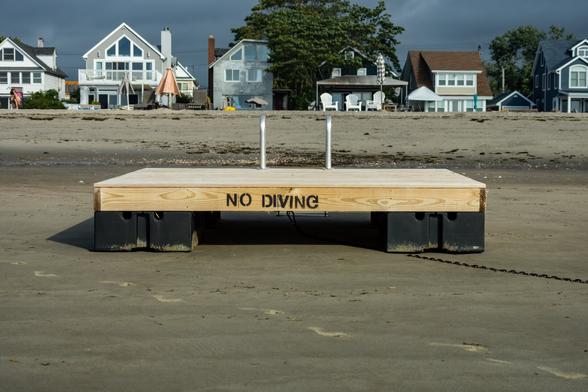 Color photograph of a square wooden floating swim dock sitting on the sands of a beach at low tide. No water is visible. Shot perpendicular to the waterline, a row of beach houses stretches across the background.

The platform itself lines up with the waterline, a metal ladder on the far side facing the houses. On the near side, the words “NO DIVING” is painted on in black, facing the camera, just above the sand.