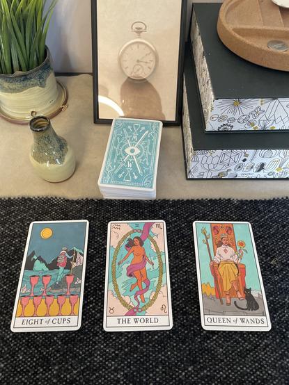 A three card draw from the modern witch tarot deck, the first card is the eight of cups with art of eight cups stacked in the foreground with a person wearing hiking gear walking toward mountains along a shoreline. The second card is the world, a nude woman with a flowing pink cloth draped around her holding two wands floating in the blue sky, bordered by a wreath of greenery. The third card is the queen of wands featuring a woman in a white blouse and yellow skirt sitting in an ornate throne holding a flaming staff in her right hand and a sunflower in her left. A black cat sits in the foreground by her left hand.