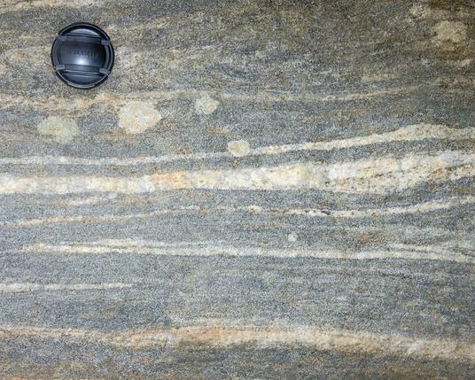 Horizontal light and dark bands of felsic and mafic minerals alternate across the on the face of this water-smoothed boulder. A 4 cm lens cap at top left provides scale.