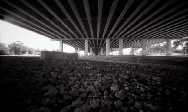 Underneath the large upper-deck of Interstate 35. Coarse gravel ground contrasts with the long smooth lines of the underside of the overpass.