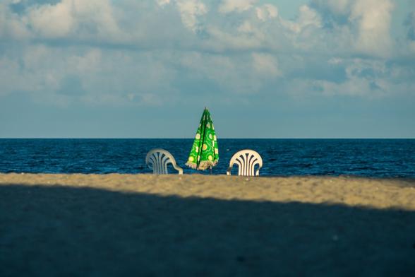 A photograph of a closed, green ,beach umbrella flanked by two white plastic chairs. In the foreground, the sand of the beach, partially covered by a shadow of a roof line, rises towards the water before falling. The chairs and umbrella are on the far side of this, so they’re cut off at the bottom - just over the “hill”. Behind them, the blue water forms a horizontal band, with a sharp horizon leading to a pale blue sky with puffy clouds.
