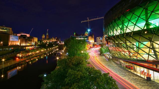 A night view of downtown Ottawa. The Rideau Canal is on the lower left of the image; Colonel By Drive, with red and yellow light trails of moving traffic, is on the lower right.

In the distance, to the left of the canal, Parliament Hill stands, illuminated; the National Arts Centre is along the canal, to the extreme left.

To the right of the canal, the peaks of the Chateau Laurier give a green glow. On the extreme right of the photo, the Shaw Convention Centre also casts green light through its windows.