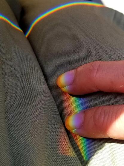 Rainbow prismatic lines on my jumpsuit. My two fingers meeting one of the rainbow bands.