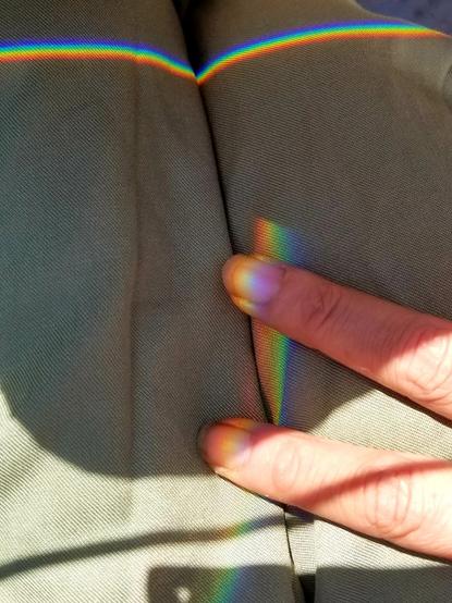 Rainbow prismatic lines on my jumpsuit. My two fingers meeting one of the rainbow bands.