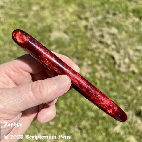 Capped pen in rich, chatoyant red material, held in maker’s hand against a green grass background.