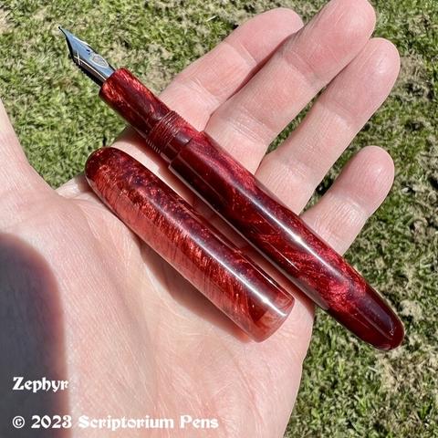 Uncapped pen in rich, chatoyant red material, held in maker’s hand against a green grass background.