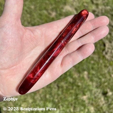 Capped pen in rich, chatoyant red material, held in maker’s hand against a green grass background.